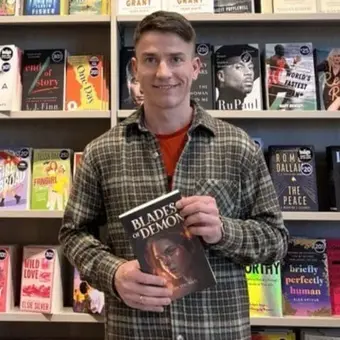 Person holding 'Blades of Demon' book in front of bookstore shelves.